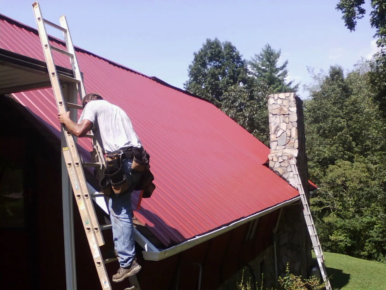 Roofer on a ladder beside a red metal roof and stone chimney — Soffit & Fascia Repair by Roofing Brothers in Mountain Dale